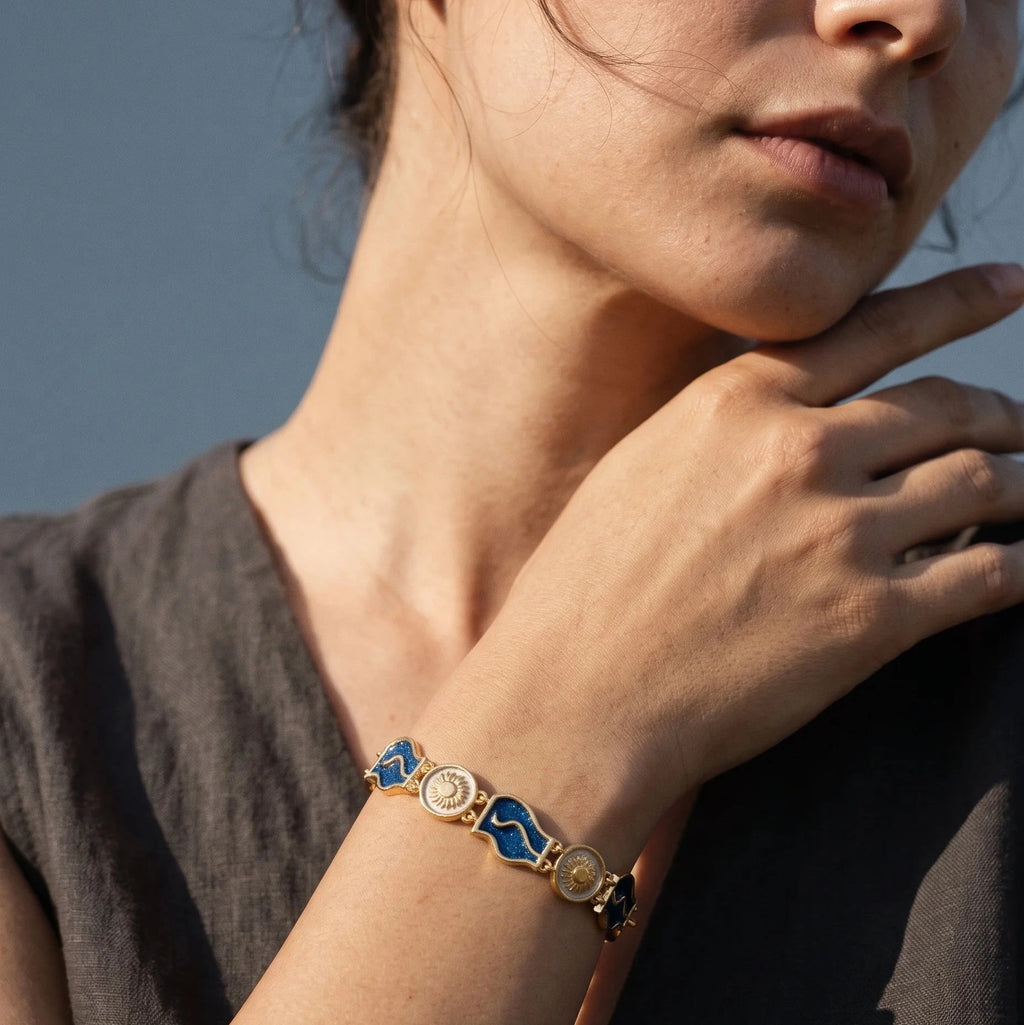 A close-up lifestyle shot of a woman wearing a gold-plated bracelet with blue and white enamel geometric links on her wrist, set against a soft blue-gray background.