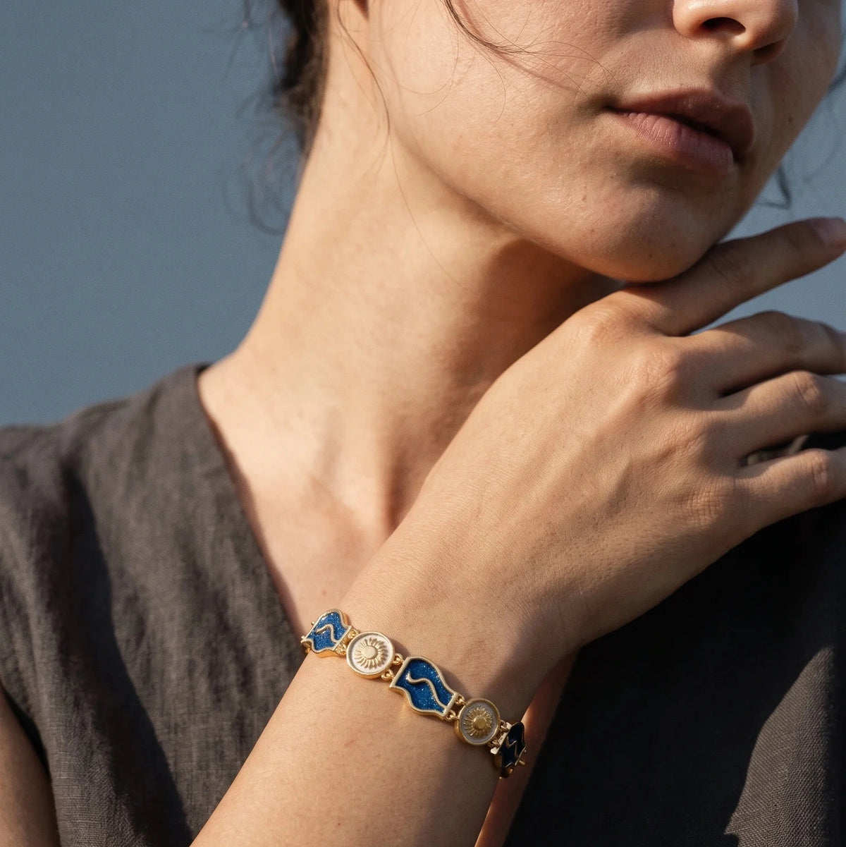 A close-up lifestyle shot of a woman wearing a gold-plated bracelet with blue and white enamel geometric links on her wrist, set against a soft blue-gray background.