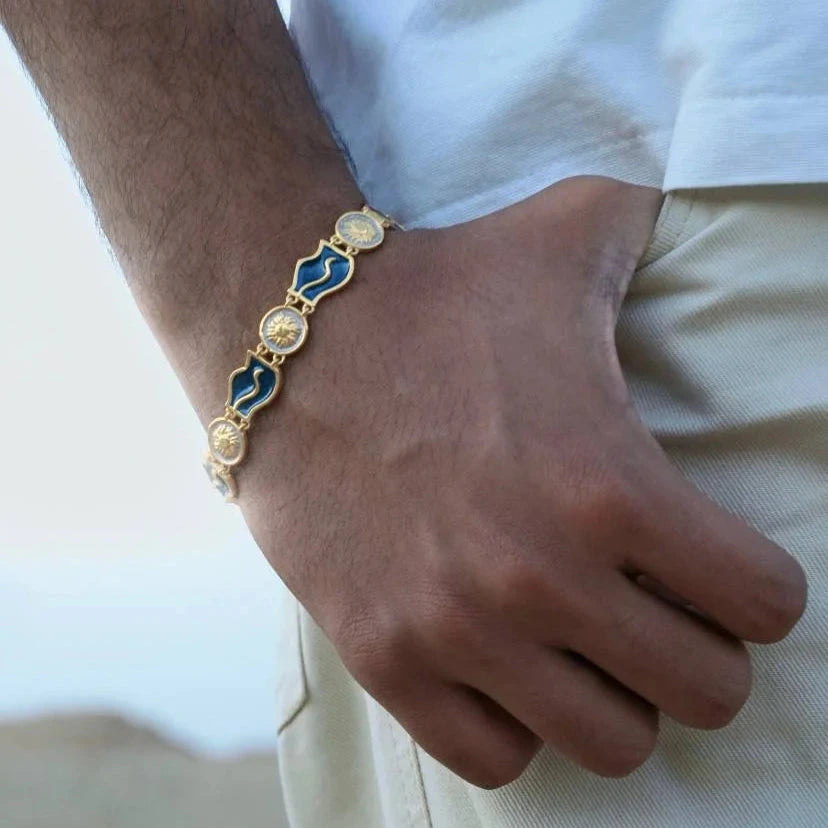 A gold chain bracelet with alternating blue and gold oval links worn on a person's wrist, shown against a light-colored shirt and pants.